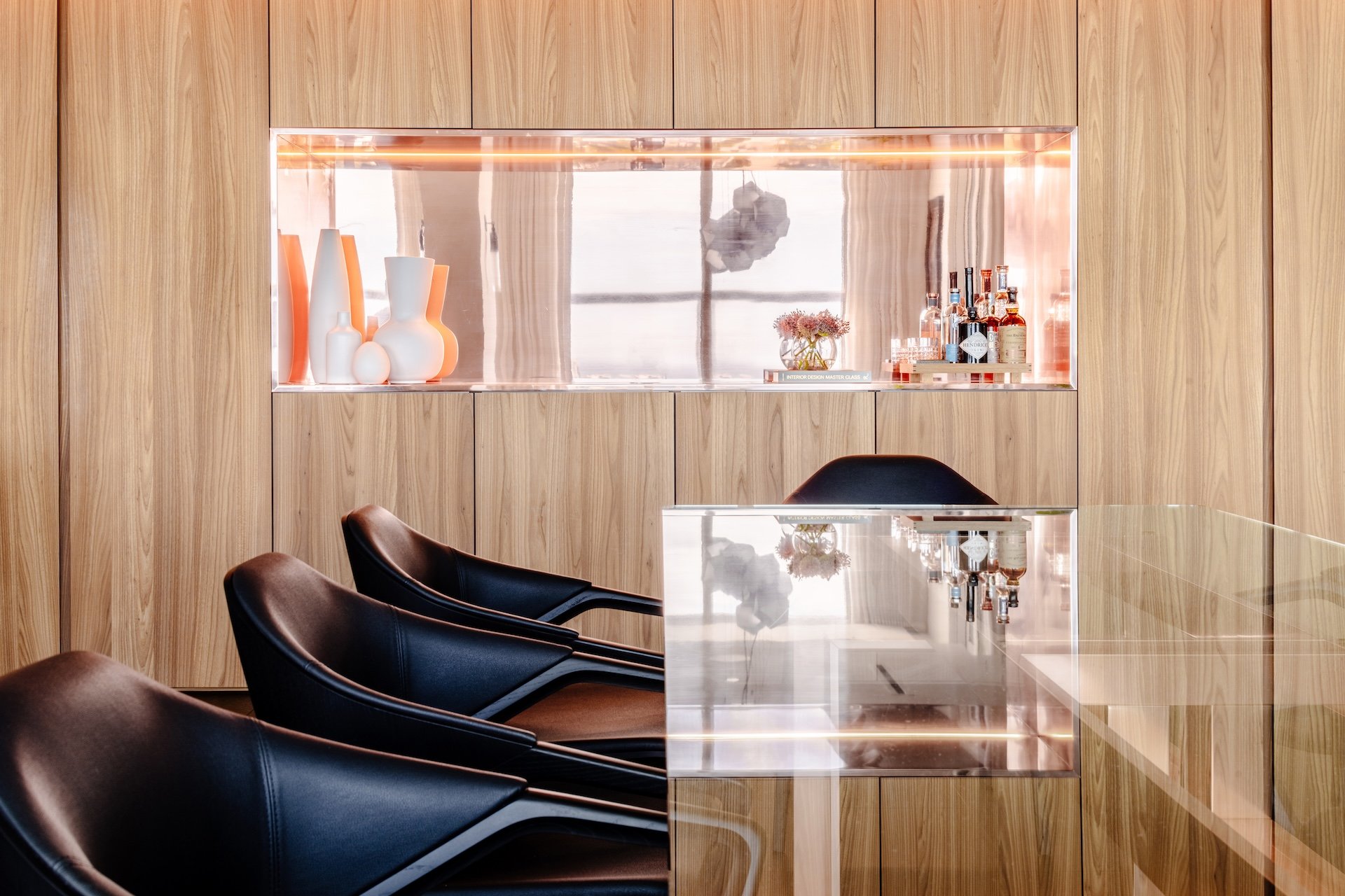 Warm wood-paneled interior with black chairs, illuminated display niche, and glass table in a modern luxury space. Warm wood-paneled interior with black chairs, illuminated display niche, and glass table in a modern luxury space.